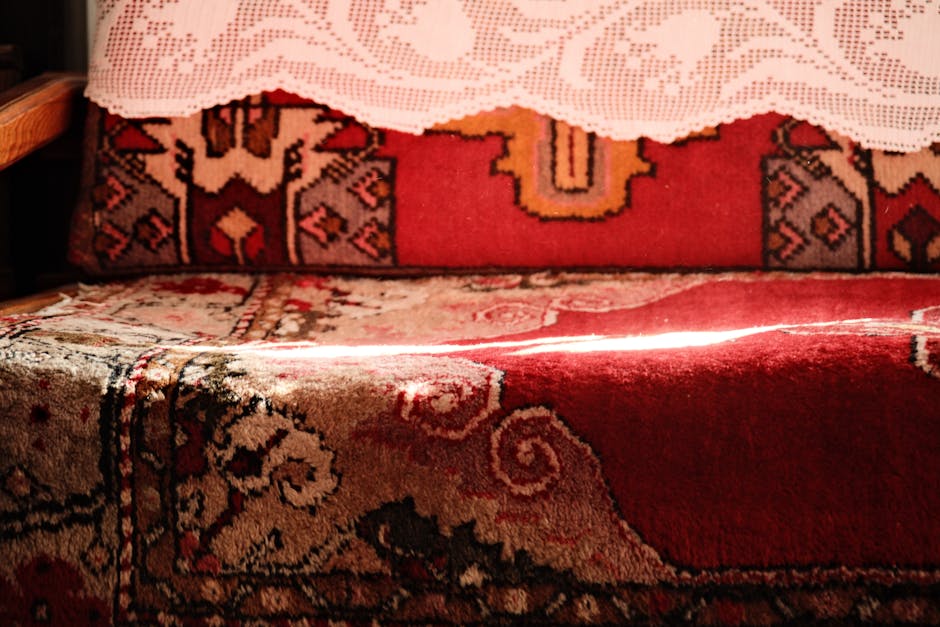 Close-up view of a traditional patterned carpet on a wooden floor in a room illuminated by natural light, partially covered by a lace tablecloth. The carpet features intricate designs in shades of red