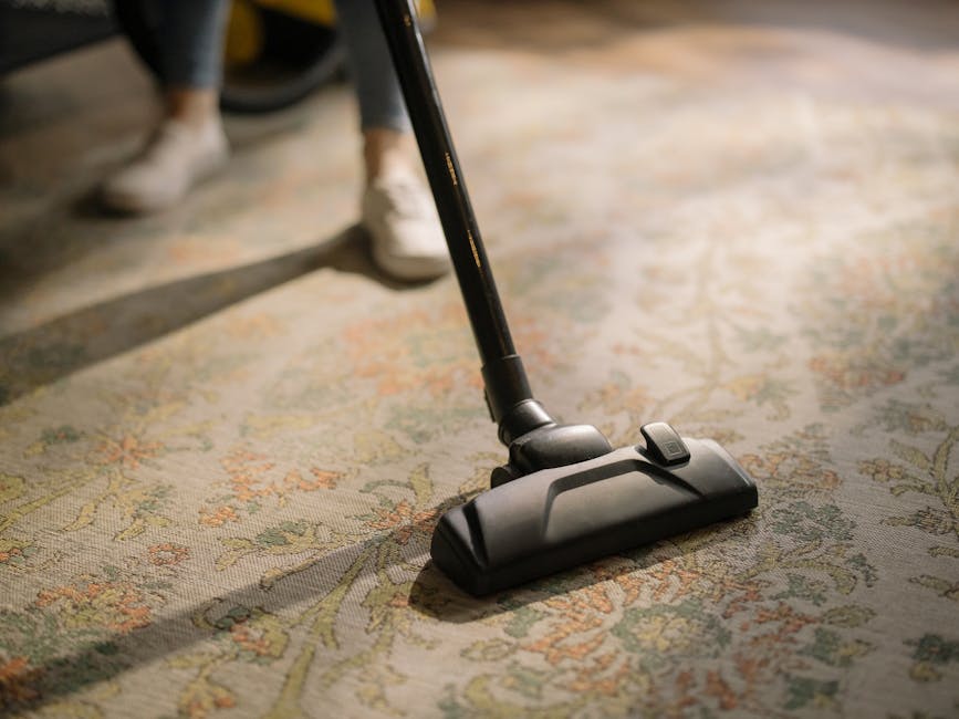 Close-up of a vacuum cleaner with a black floor attachment being used to clean a patterned area rug in a well-lit room, with a person wearing white shoes and dark pants standing nearby. The rug featur
